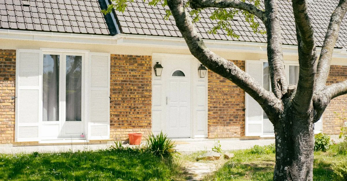 Vue extérieure ensoleillée d'une maison en briques avec un arbre et un chemin de jardin luxuriant.