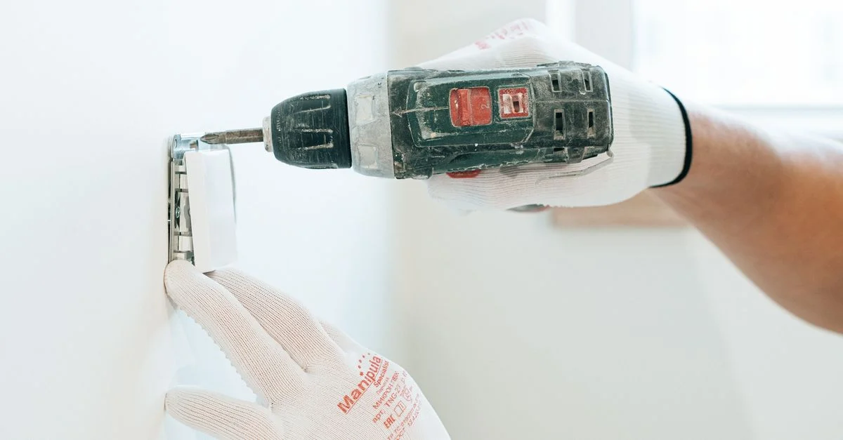 Des mains gantées utilisant une perceuse pour installer une prise électrique sur un mur, en mettant l'accent sur la précision et la sécurité.