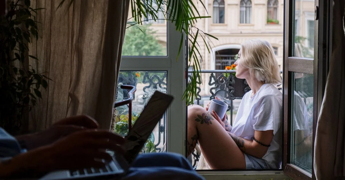 Jeune femme profite d'un moment paisible avec un café sur un balcon, ordinateur portable ouvert à l'intérieur.