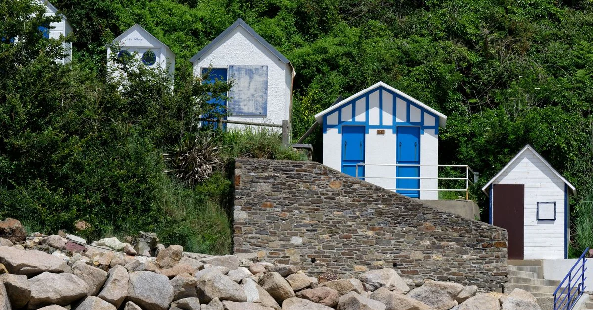 Cabanes de plage colorées avec mur en pierre et verdure à Barneville-Carteret, France.