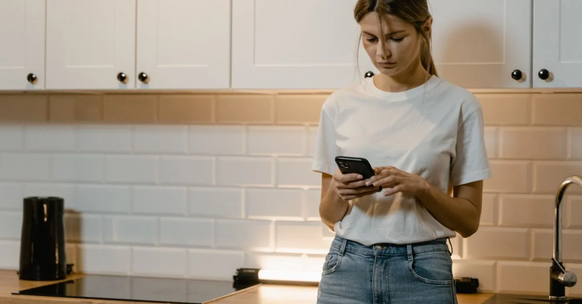 Jeune femme dans une cuisine utilisant un smartphone, avec une expression concentrée et réfléchie.
